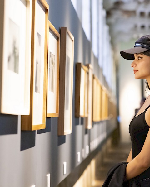 A person wearing a black tank top and cap is looking at framed artworks displayed on a gallery wall.