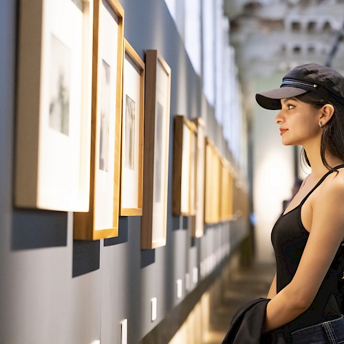 A person wearing a black tank top and cap is looking at framed artworks displayed on a gallery wall.