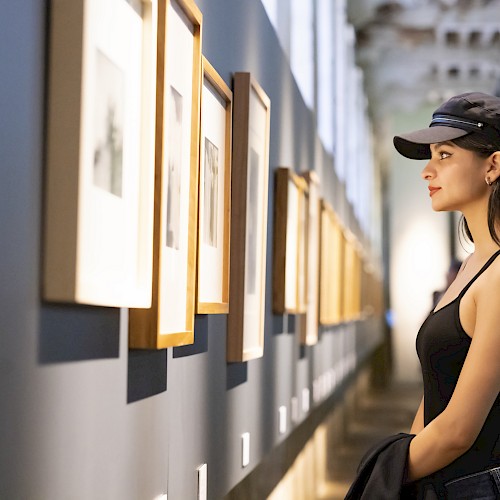 A woman wearing a cap looks at framed artworks on a gallery wall, with a blurred background and soft lighting enhancing the scene.