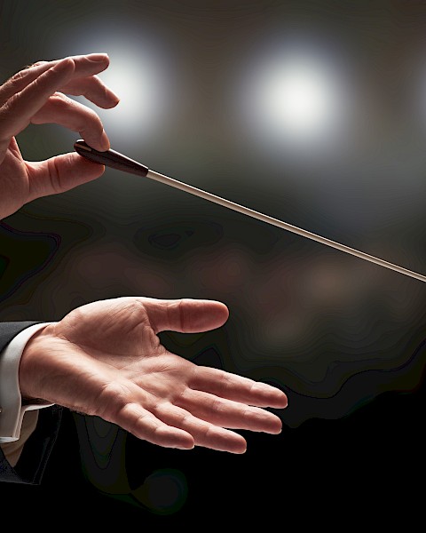 A conductor's hands are holding a baton against a blurred background of lights, suggesting a musical performance setting.