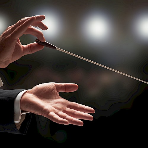 A conductor's hands are holding a baton against a blurred background of lights, suggesting a musical performance setting.