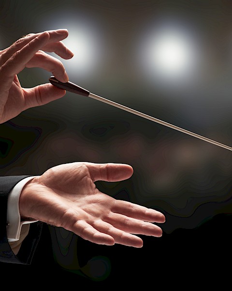 The image shows a conductor's hands holding a baton in front of blurred lights, suggesting an orchestral setting.