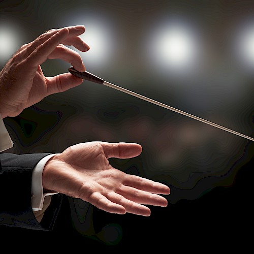 The image shows a conductor's hands holding a baton in front of blurred lights, suggesting an orchestral setting.