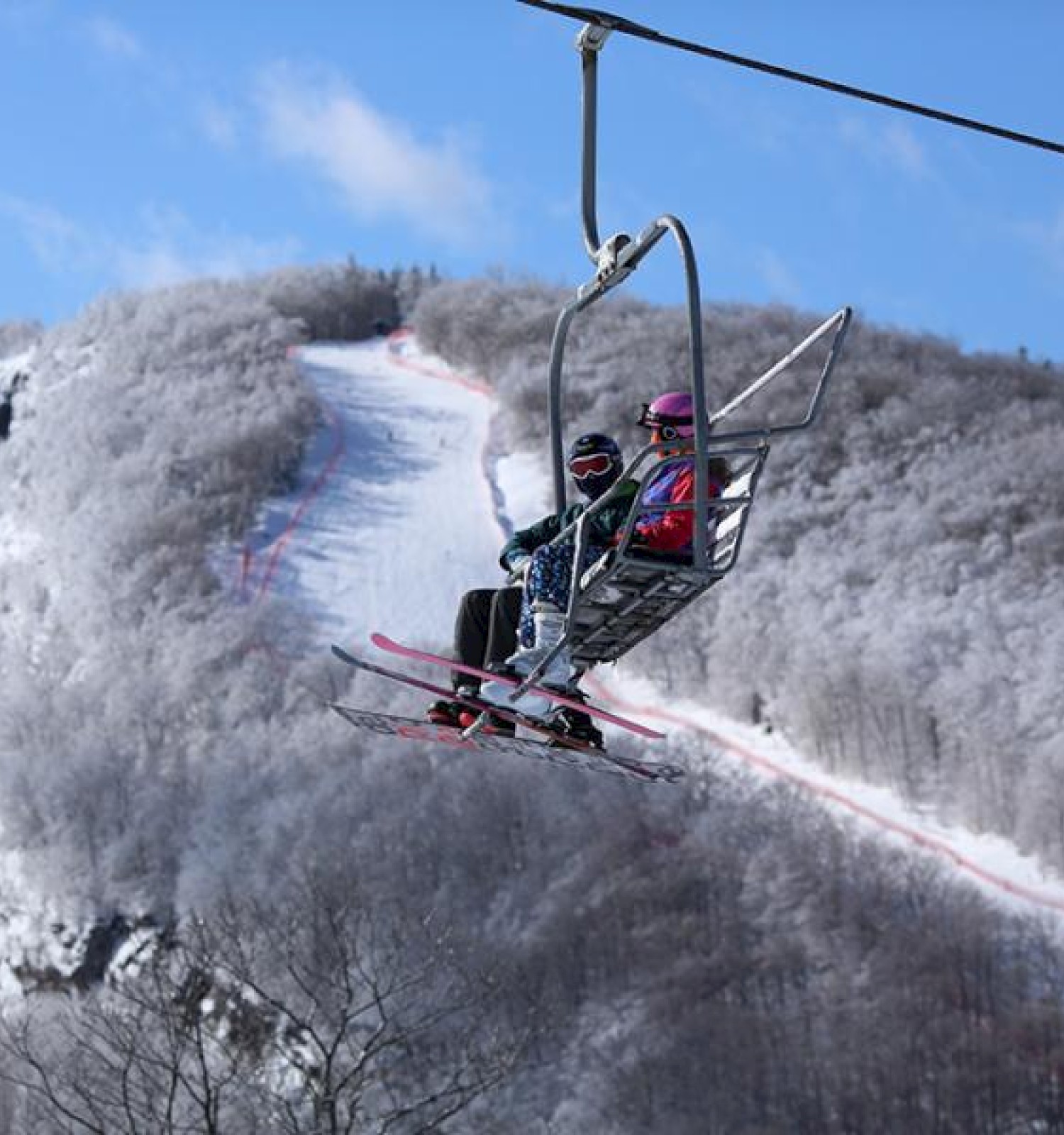 Two people on a ski lift ride over a snowy mountain landscape, with a clear blue sky in the background.