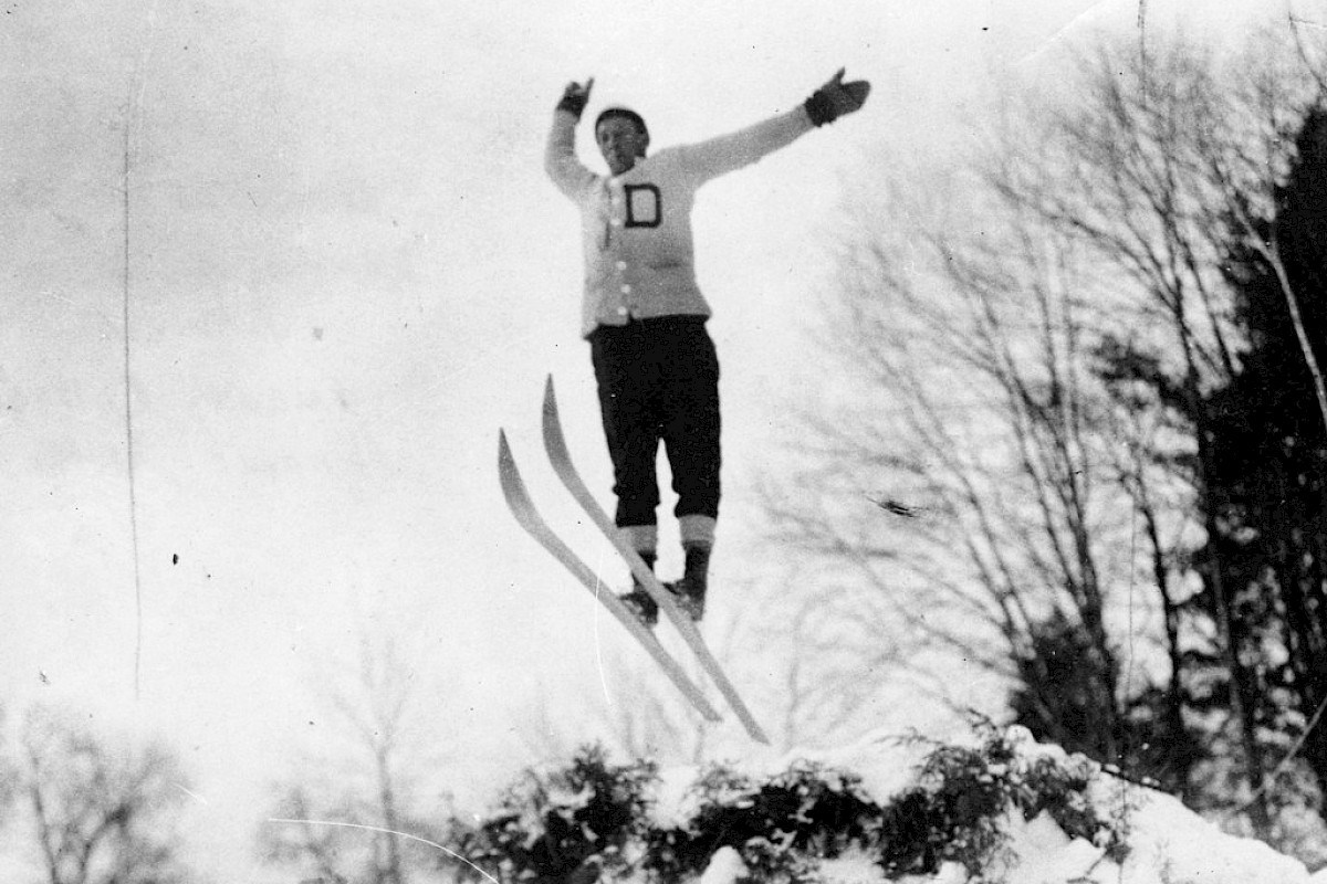 A person wearing a lettered cardigan is mid-air on skis, performing a jump with arms raised, snowy slope and bare trees in the background.