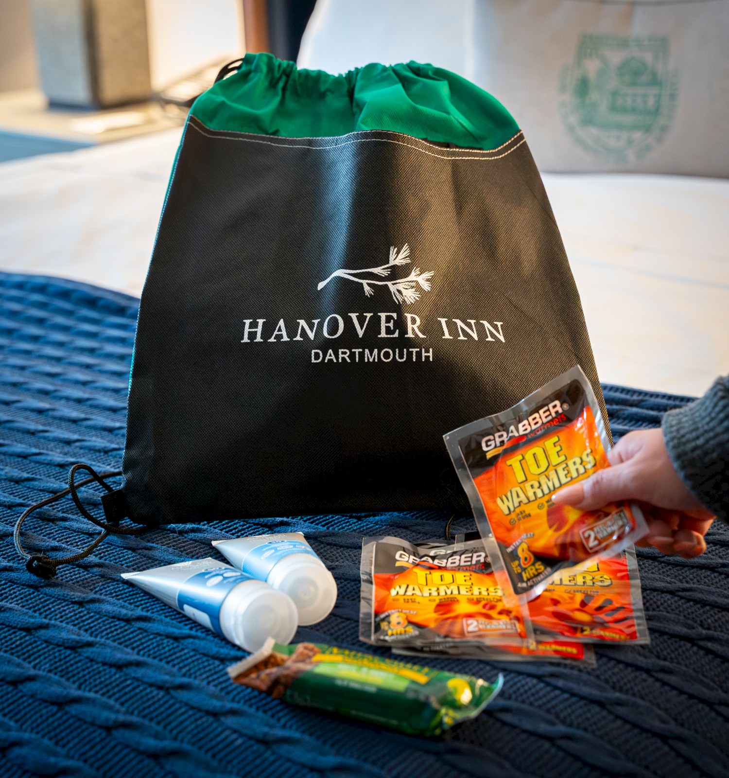 A Hanover Inn tote bag with hair ties, packets of snacks, and a small bottle, arranged on a blue tablecloth.