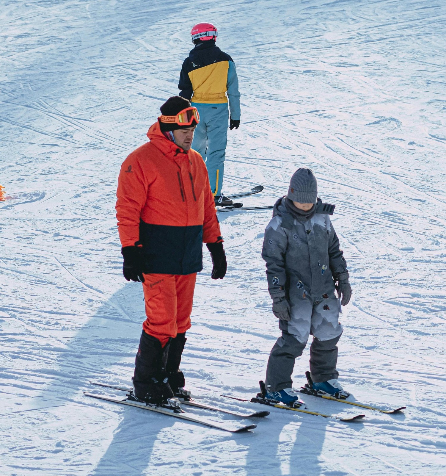 People skiing on a snowy slope: a child in gray gear learns alongside an adult in bright red, with others in the background near a cone.