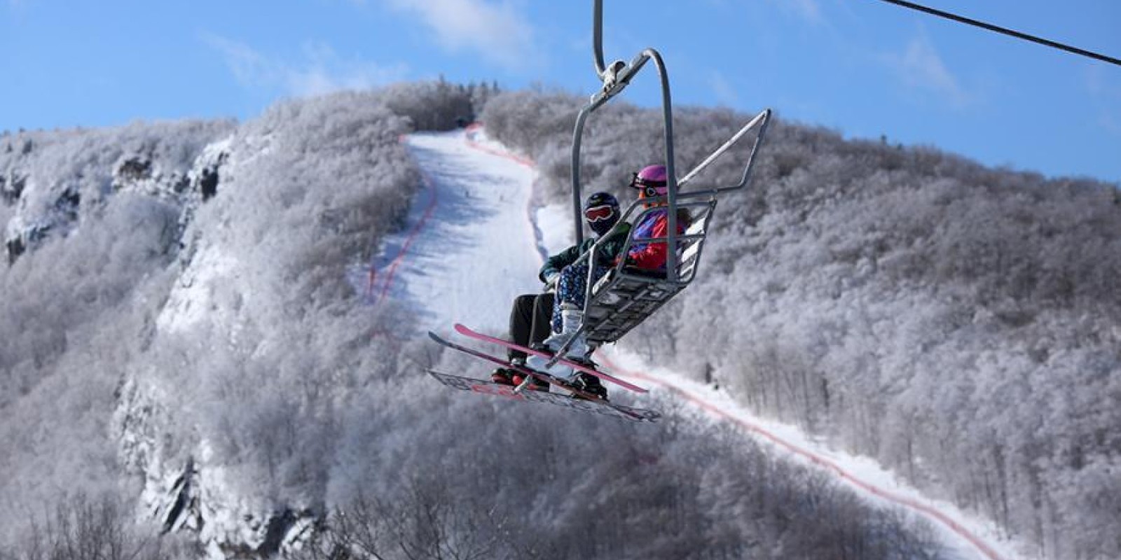 Two people on a ski lift ascending a snowy mountain with a ski trail visible in the background under a bright blue sky.