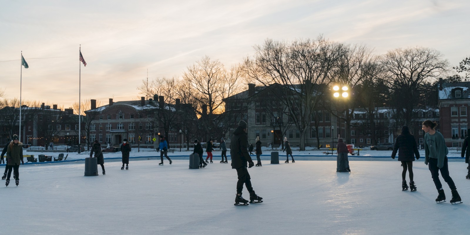 People are ice skating on an outdoor rink at dusk with surrounding buildings and trees.