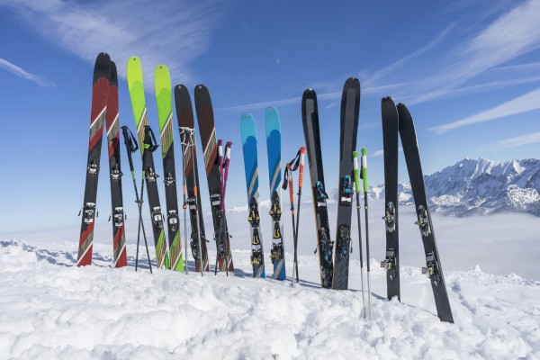 A row of colorful skis and poles stands upright in the snow against a mountain backdrop and blue sky.