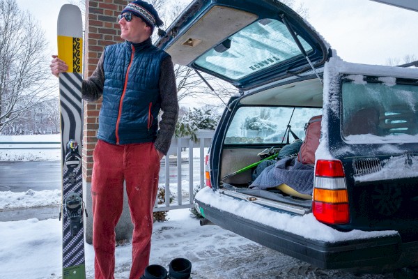 A person in winter attire stands by a car holding skis. Snowy ground, ski boots nearby, car hatch open with snowy surroundings.