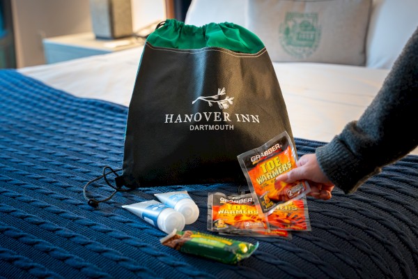 A Hanover Inn bag on a bed with toiletries, toe warmers, and a snack bar nearby, as someone reaches for a toe warmer.