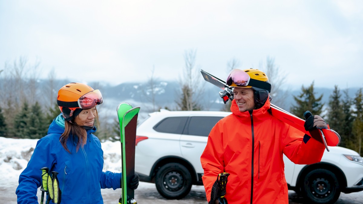 Two people in winter gear hold skis and smile near a vehicle, with a snowy, mountainous background.