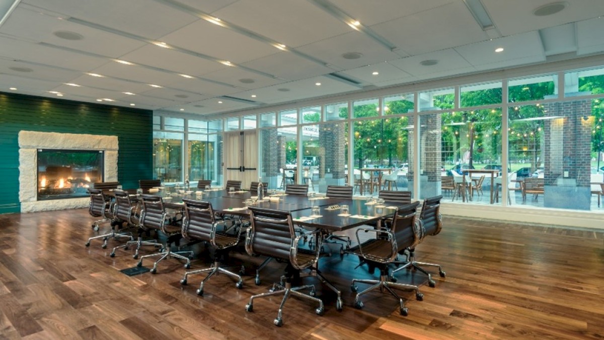 A modern conference room with a large table, chairs, a fireplace, and glass windows showing a patio with outdoor seating.