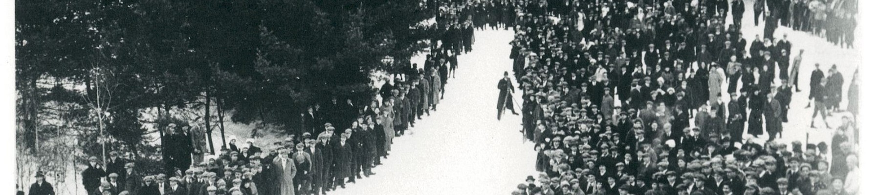 A large group of people standing in snow, lined up along a road, surrounded by trees, creating a striking visual pattern in the photograph.