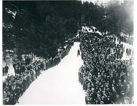 A large group of people standing in snow, lined up along a road, surrounded by trees, creating a striking visual pattern in the photograph.