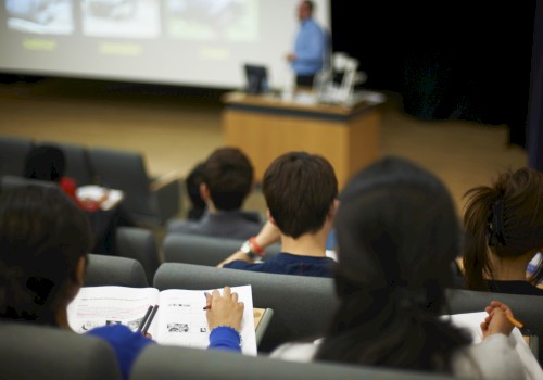 People sitting in a lecture hall, taking notes, and watching a presentation on a screen.