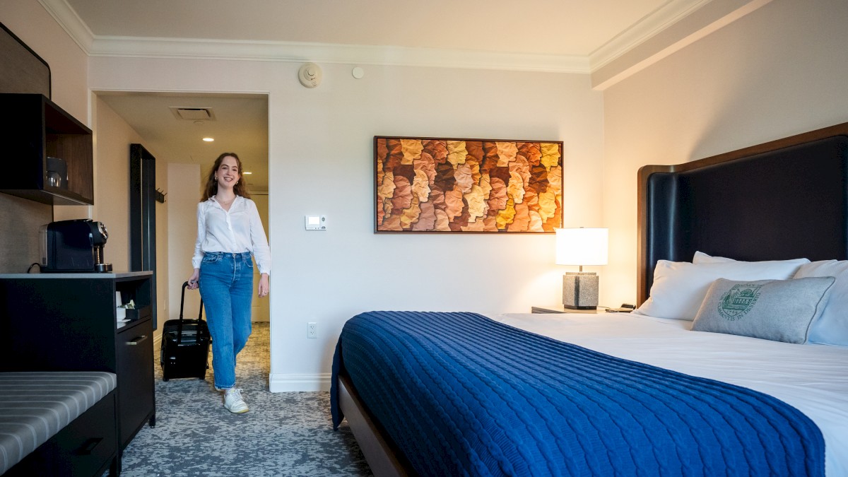 A person enters a hotel room with a neatly made bed, blue bedding, artwork on the wall, and a suitcase in hand.