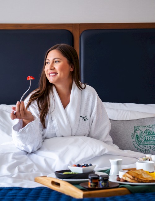 A person in a white robe enjoys breakfast in bed, holding a fork with fruit. There's a tray with breakfast items and drinks.