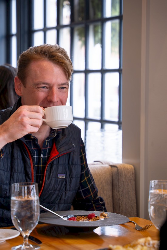 A man is enjoying coffee and breakfast in a cozy cafe with large windows and other patrons in the background.