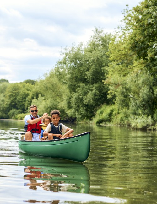 A man and two children are paddling in a green canoe on a river surrounded by lush greenery under a cloudy sky.