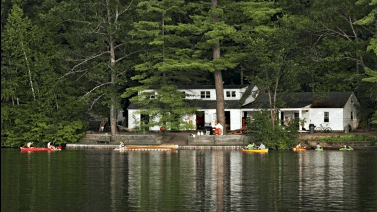 A lakeside house surrounded by trees, with people in kayaks and canoes on the water in front.
