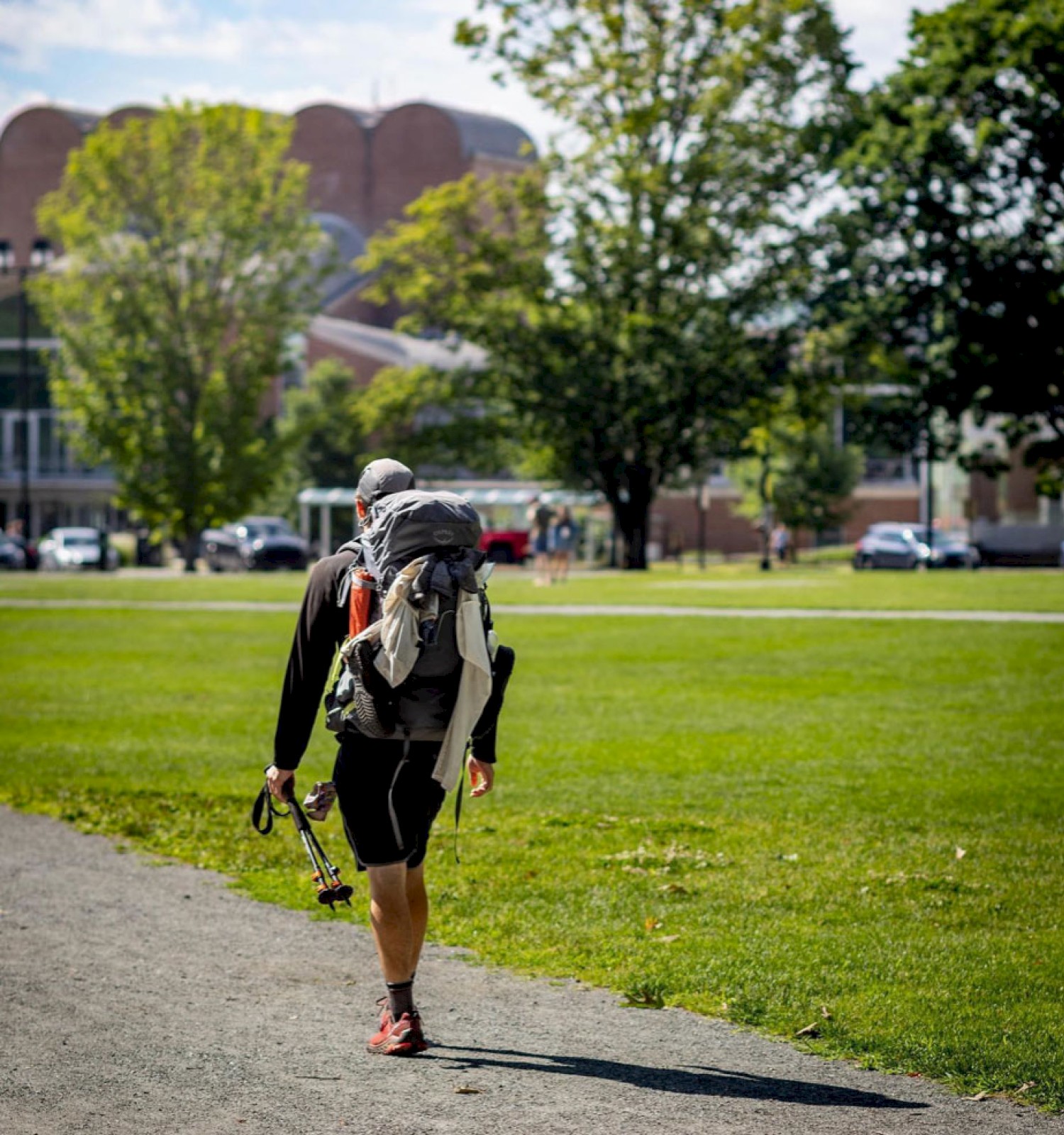 A person with a backpack walks across a grassy area towards buildings, carrying hiking poles. Trees and parked cars are visible in the background.