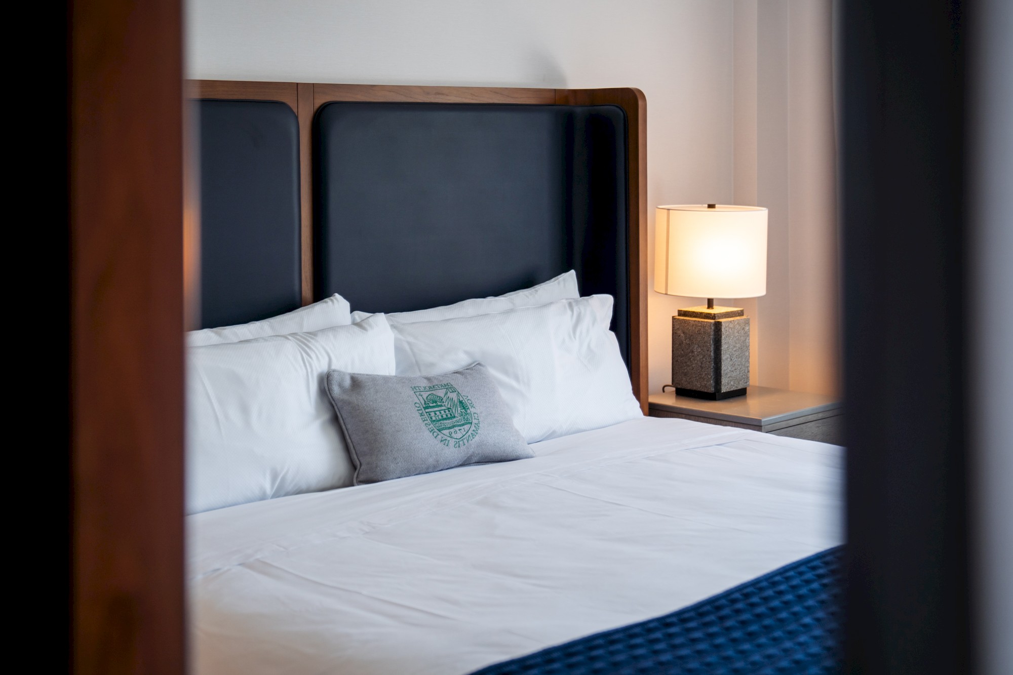A neatly made bed with white pillows, a decorative cushion, a dark headboard, and a side table with a lit lamp is visible in the image.