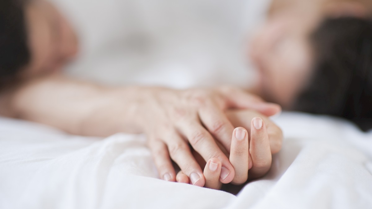 Two people laying on a bed, holding hands gently, with a soft focus on the scene creating a warm and intimate atmosphere.