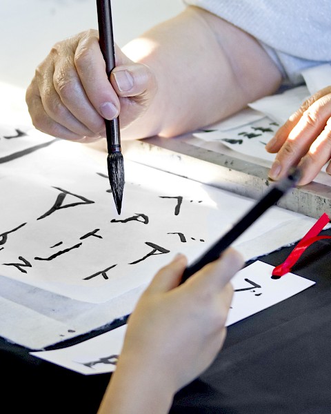 People practicing calligraphy with brushes and ink, creating text on white paper, focusing on traditional writing techniques.