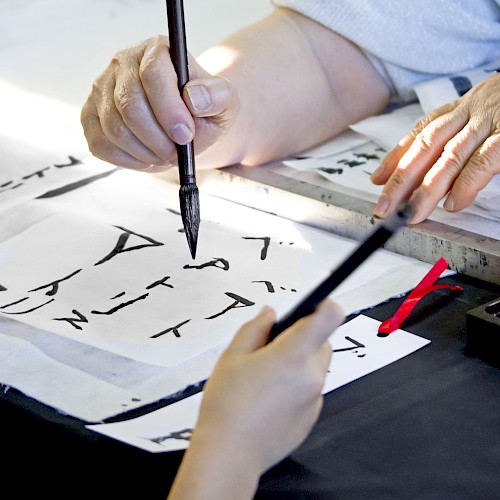 People practicing calligraphy with brushes and ink, creating text on white paper, focusing on traditional writing techniques.