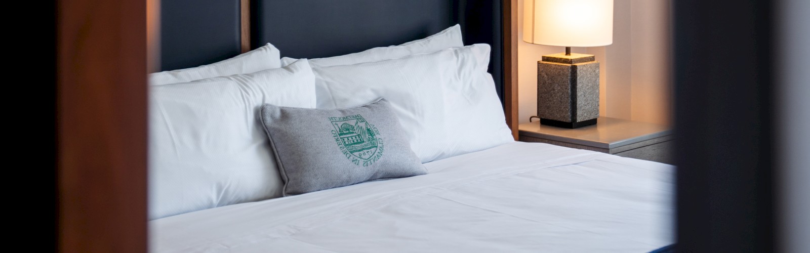 A neatly made bed with white pillows, a decorative cushion, a dark headboard, and a side table with a lit lamp is visible in the image.