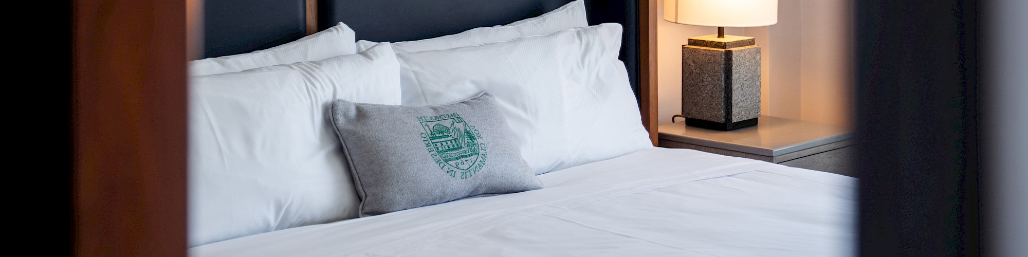 A neatly made bed with white pillows, a decorative cushion, a dark headboard, and a side table with a lit lamp is visible in the image.