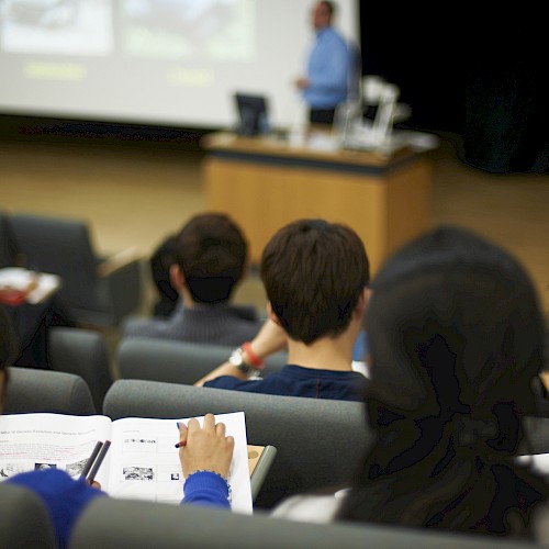 Students are sitting in a lecture hall, watching a presentation. A presenter stands at the front near a screen.