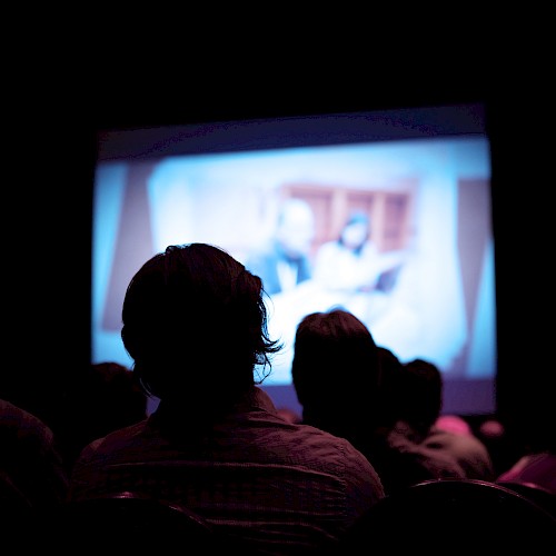 A group of people are watching a blurry projection in a dark room, indicating a movie or presentation.