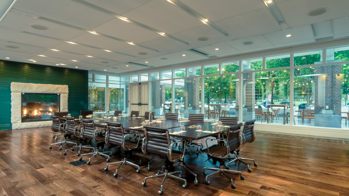 A modern conference room with a large table, many chairs, a fireplace, and a window wall offering a view of the outdoors, well-lit and spacious.