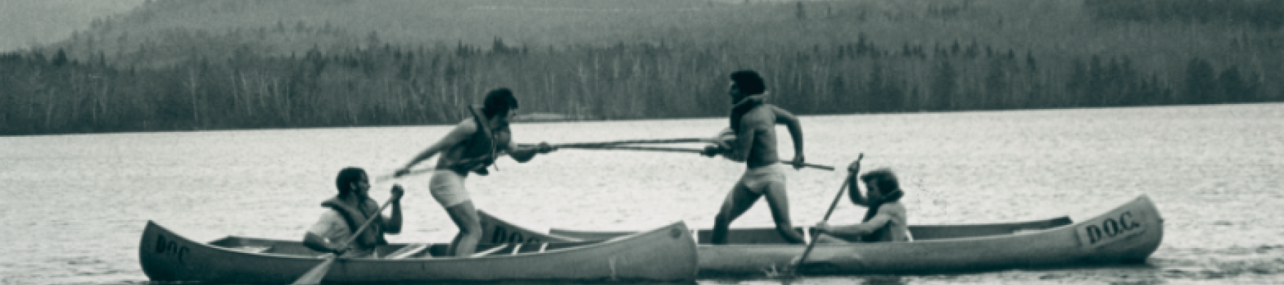 Four people in two canoes seem to be jousting on a lake, surrounded by trees and hills in the background.