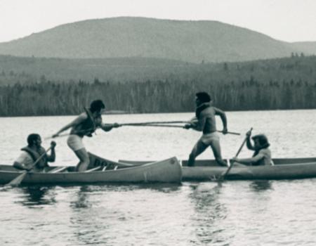Four people in two canoes seem to be jousting on a lake, surrounded by trees and hills in the background.