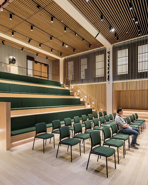 A modern lecture hall with tiered seating, several green chairs, and two people talking near the entrance.