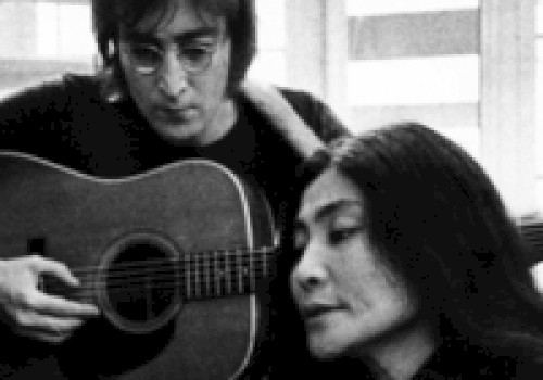 A person with glasses playing an acoustic guitar, sitting next to another person with long hair, indoors with large windows in the background.