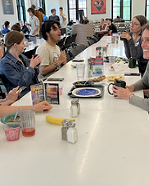 People are sitting at a long table in a cafeteria, eating and talking. Various dishes are on the table, including a banana and drinks.