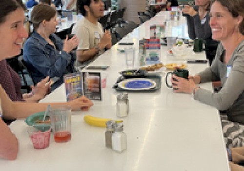 People are sitting at a long table in a cafeteria, eating and talking. Various dishes are on the table, including a banana and drinks.