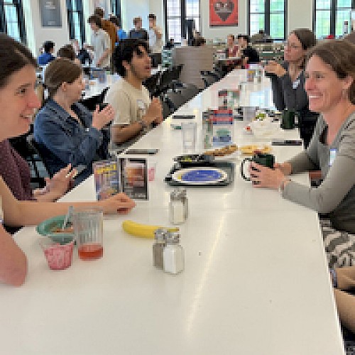 People are sitting at a long table in a cafeteria, eating and talking. Various dishes are on the table, including a banana and drinks.