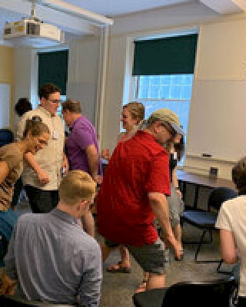 A group of people, in casual clothing, are indoors playing a game of musical chairs, moving around closely arranged chairs.