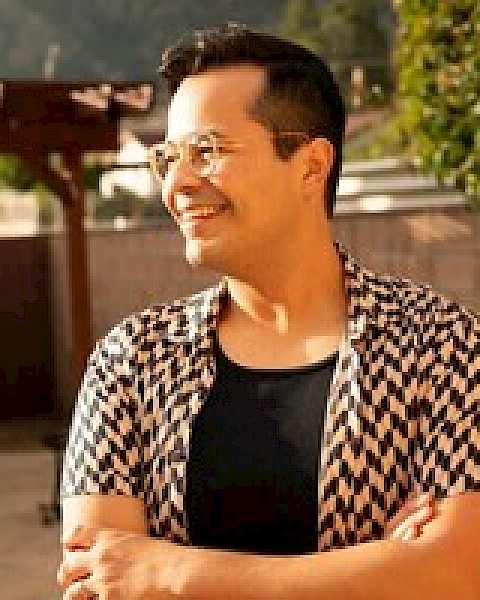 A person with glasses, wearing a patterned shirt, standing outdoors and smiling with arms crossed.