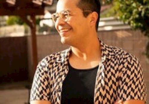 A person with glasses, wearing a patterned shirt, standing outdoors and smiling with arms crossed.