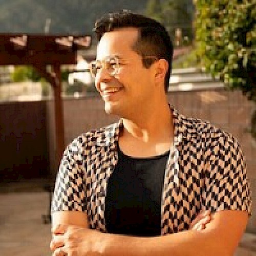 A person with glasses, wearing a patterned shirt, standing outdoors and smiling with arms crossed.
