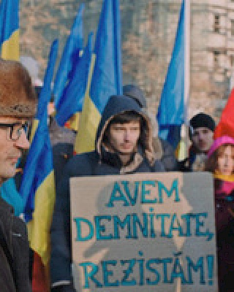 A group of people are holding flags and a sign in a protest. The sign reads, 