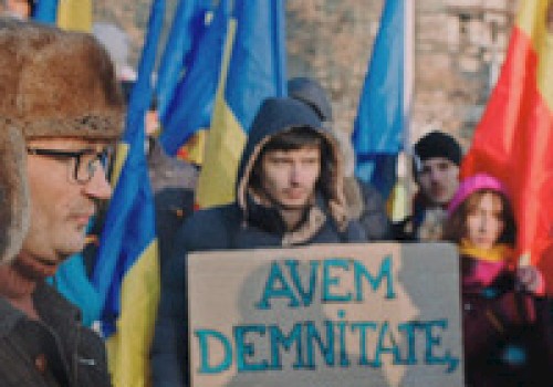 A group of people are holding flags and a sign in a protest. The sign reads, 