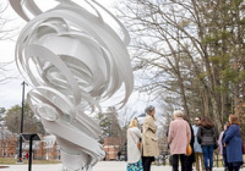 A group of people stands near a white abstract sculpture outside, surrounded by trees on a cloudy day.